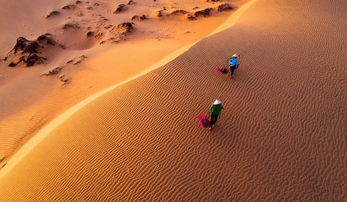 Hai người đội nón lá gánh hàng đi trên đồi cát vàng nhìn từ trên cao tại khu vực Mũi Né