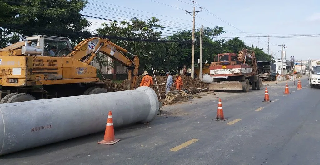 Công nhân và máy xúc thi công cạnh ống cống bê tông lớn trên mặt đường, có cọc tiêu giao thông
